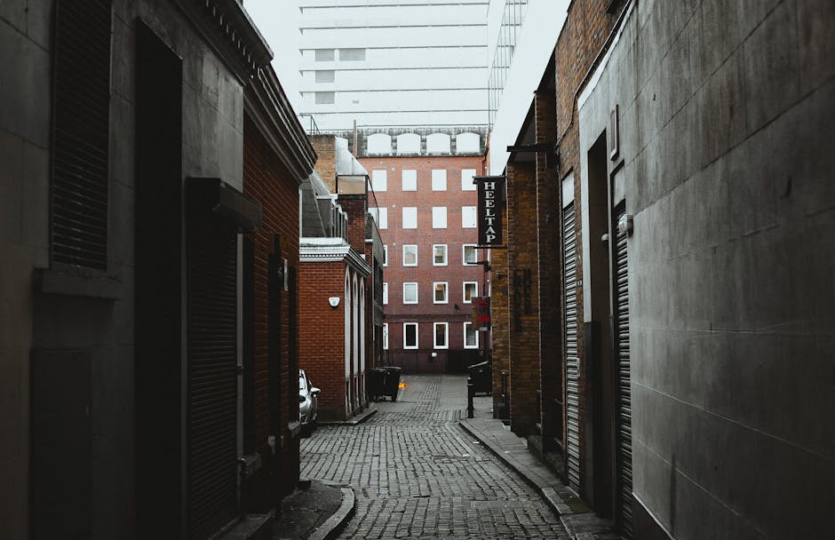 A narrow urban alleyway showing mixed building facades with brick and concrete finishes. The foreground features dark-colored rolling shutters on the left and right sides of the alley, with the left s